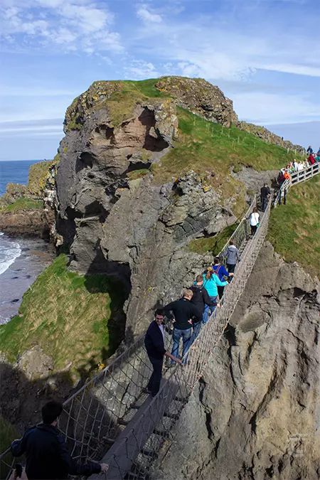 People crossing Carrck-a-Rede Rope Bridge on a sunny day, with cliffs and shoreline visible beyond.