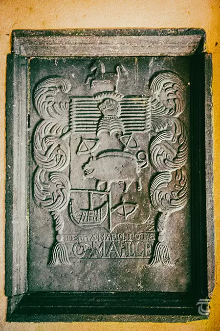 A plaque bearing the coat of arms for the O MAILLE (O'Malley family). A large boar is depicted in its centre, and it also shows three bows, a boat, and a prancing horse.