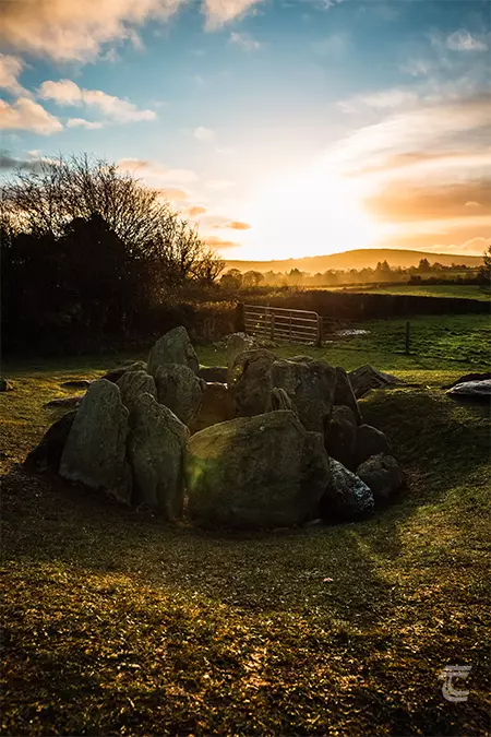 The passage tomb stands in a field bounded by a gateway. Sun setting on the horizon.